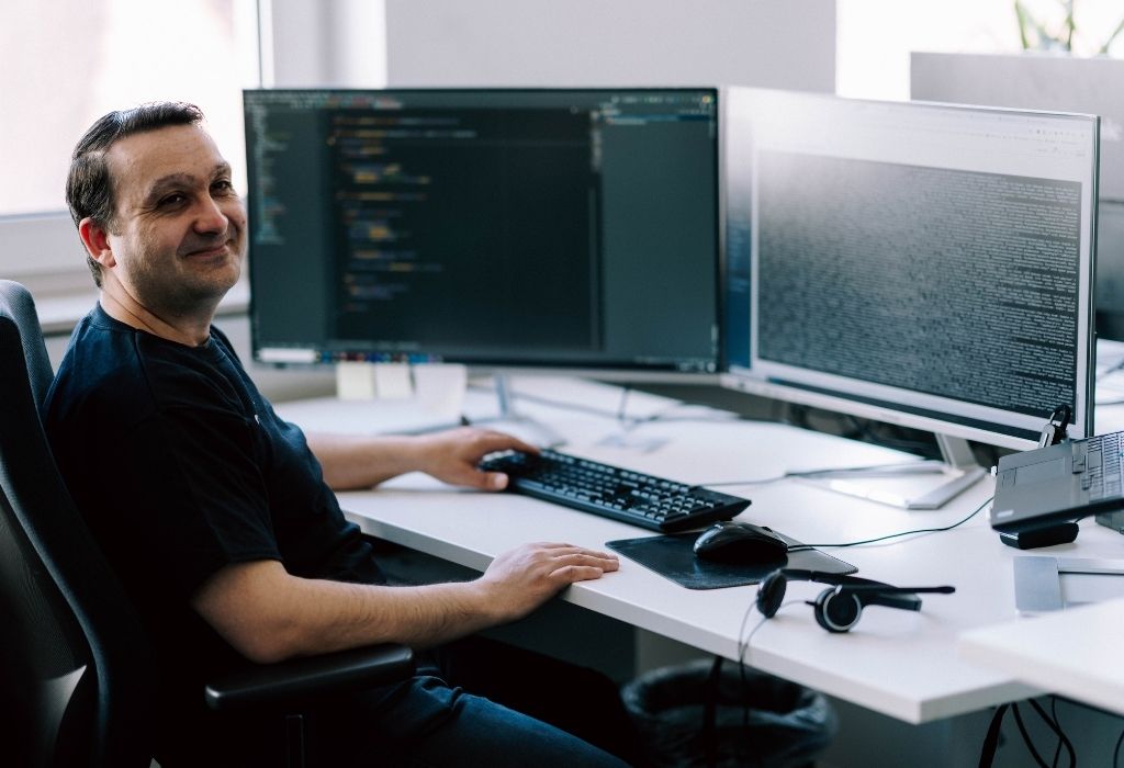 Satelliten-Blogseiten Content-Bilder (1) Freundlich lächelnder Mann sitzt an einem modernen Arbeitsplatz vor mehreren Monitoren im Büro und steht sinnbildlich für professionelle Magento-Beratung und technische E-Commerce-Expertise