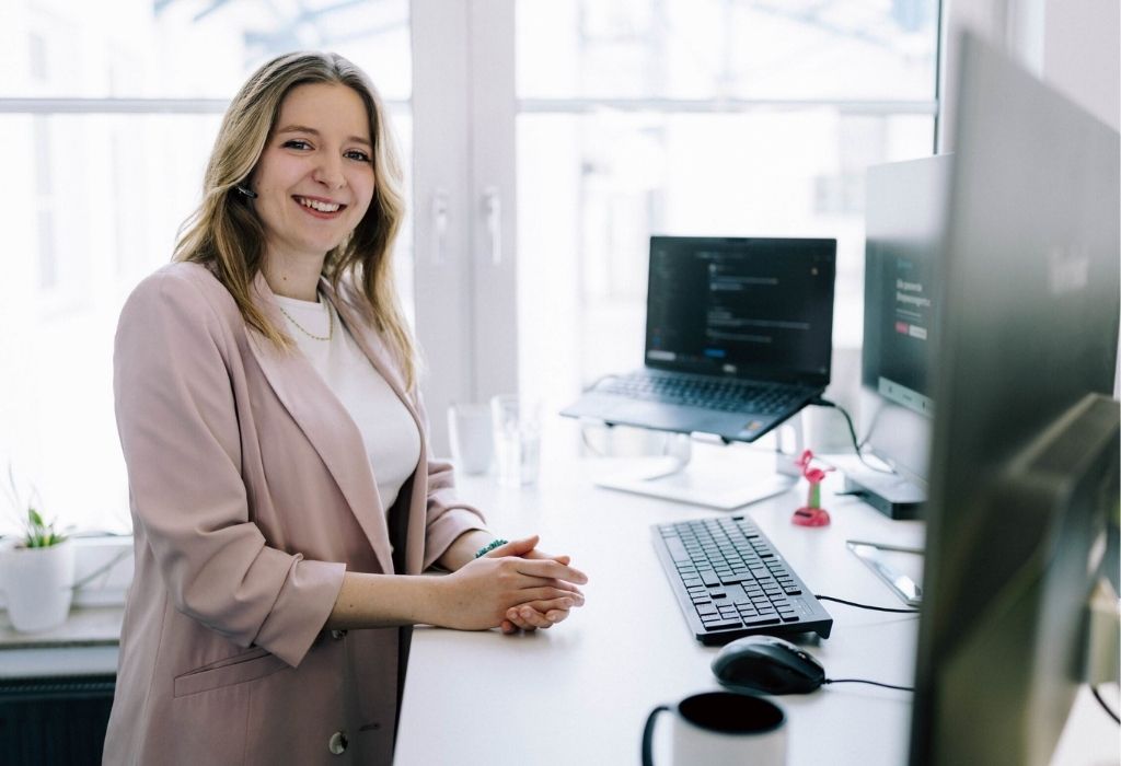 Satelliten-Blogseiten Content-Bilder (6) Freundlich lächelnde Frau sitzt an einem modernen Schreibtisch vor einem Computerbildschirm in einem hellen Büro und symbolisiert professionelle Magento-Beratung für E-Commerce-Projekte