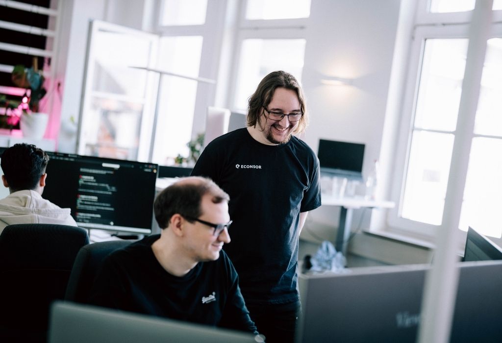 Webentwicklung-heilderberg Zwei Entwickler lächeln und diskutieren gemeinsam an ihren Schreibtischen, während sie an einem Webentwicklungsprojekt arbeiten.